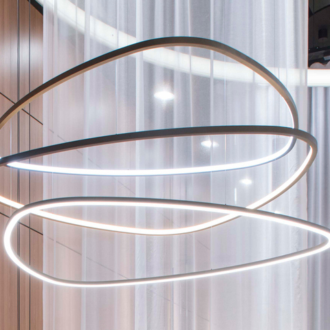 In the foreground, three narrow ring lights in the shape of a rounded triangle are suspended a short distance above each other, with the lowest one shining directly downwards and the two upper ones shining into the centre of the lights. In the background, a brown wooden wall and a slightly transparent white curtain, and a modern TOCCATA ring chandelier.