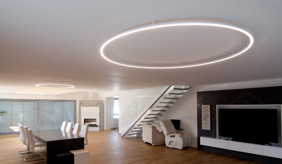 Ringleuchte SLIM DIRECT als Deckenanbauleuchte (Montageart G - Technikeinheit in Zwischendecke) Großzügiger Wohnbereich mit Esstisch und Kamin in Weiß-Schwarz-Tönen gehalten. An der Decke zwei direkt strahlende Ringleuchten, SLIM DIRECT, die mit kleinen Deckenabstandhaltern an die Decke montiert sind. Das Zuleitungskabel führt in der Nähe eines Abstandhalters in die Decke.
