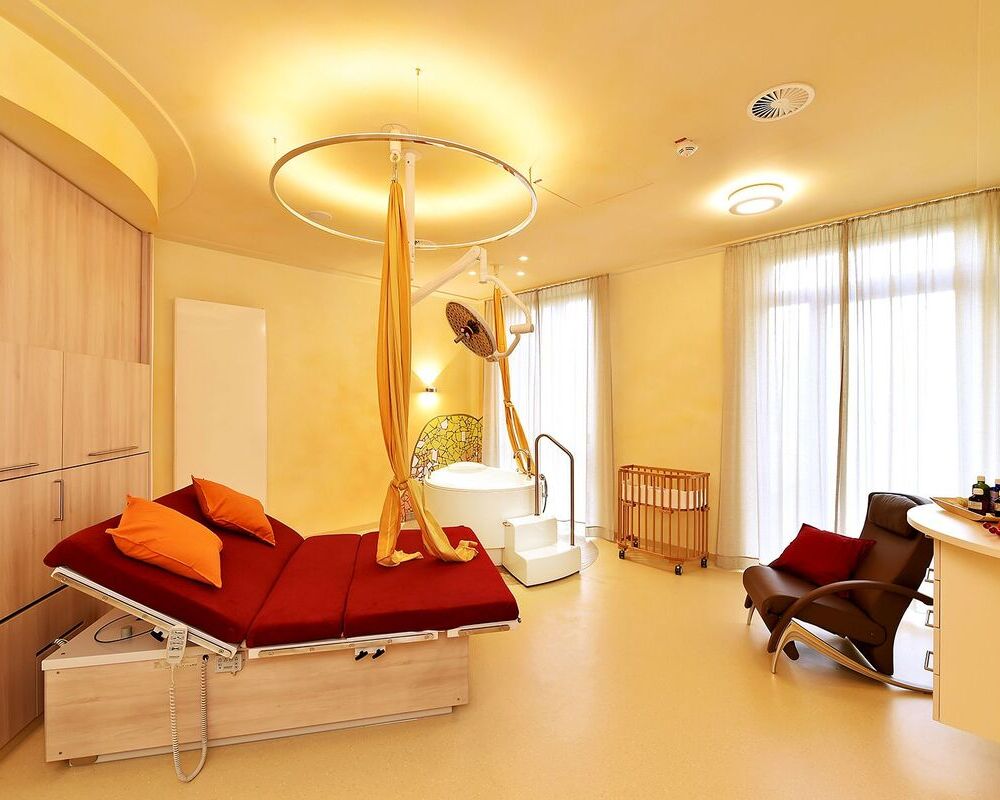 Slim pendant light SLIM INDIRECT creates ring-shaped indirect light Modern, cosy delivery room at St. Josef's Hospital in Dortmund with yellow walls and a wooden wall behind a wide red bed, above which hangs a slender round ring light with a polished aluminium housing, SLIM INDIRECT. The lamp provides indirect lighting, illuminating the ceiling and creating a ring-shaped circle of light.