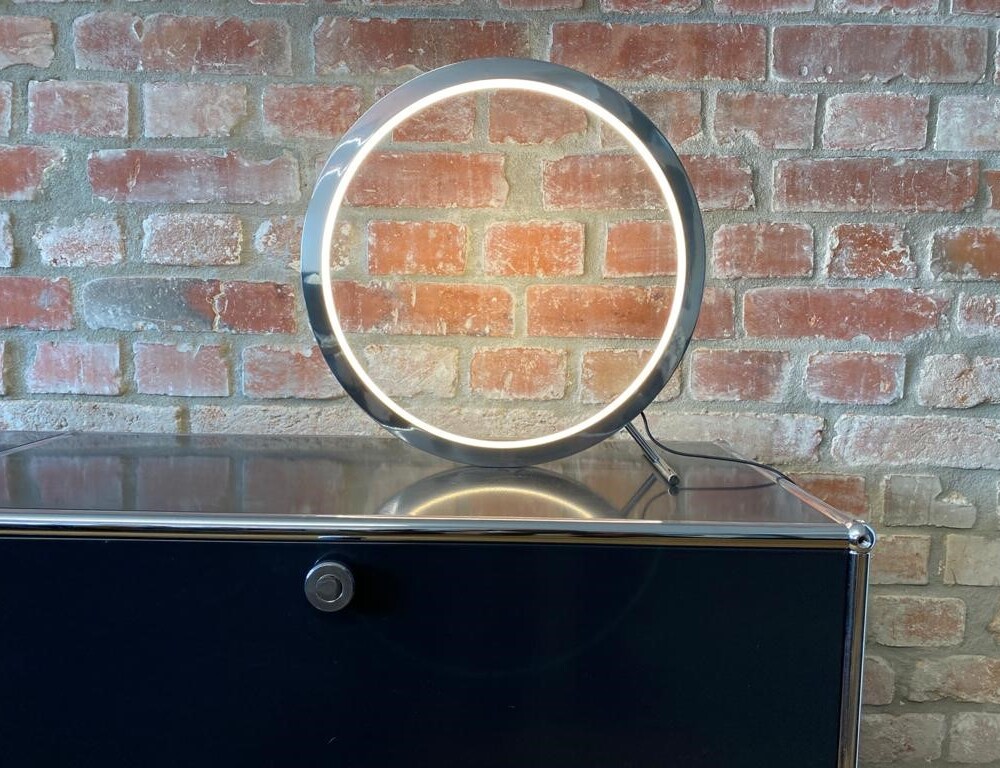 Tischleuchte wie ein aufgestellter Ring, der nach innen leuchtet aus Aluminium poliert, SLIM-TABLE, auf einem schwarzen Sideboard vor einer Backsteinwand.