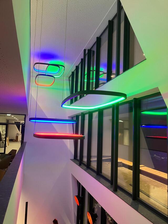 Innenraum eines modernen Gebäudes, links zeigt es einen Gang in höherem Stock, im Atrium/Treppenhaus, dass sich zu der Glasaußenfassade ergibt sind 6 großformatige viereckige, SOFT QUADRA, und rechteckige, SOFT SQUARE, Rahmenleuchten mit RGB in unterschiedlichen Höhen abgehangen, jede erstrahlt in unterschiedlicher Farbe: rot, grün, blau.