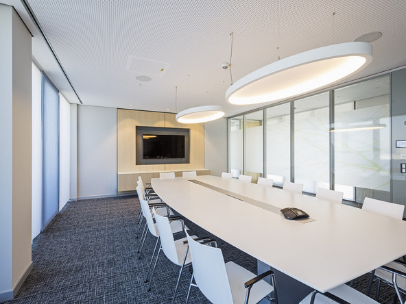 A light-coloured meeting room with a rectangular white table and white chairs in front of a TV screen. Two large elliptical white acoustic lights, SOFT ELLIPSE L+A, are suspended above the table. | © Eckhart Matthäus | www.em-foto.de