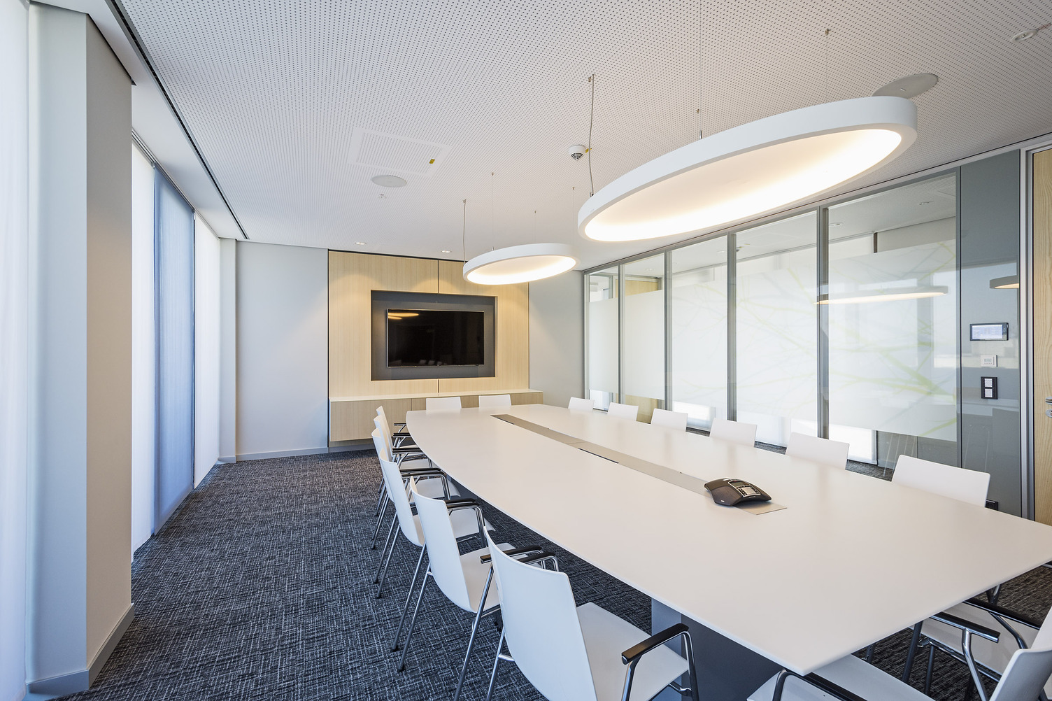 A light-coloured meeting room with a rectangular white table and white chairs in front of a TV screen. Two large elliptical white acoustic lights, SOFT ELLIPSE L+A, are suspended above the table.
