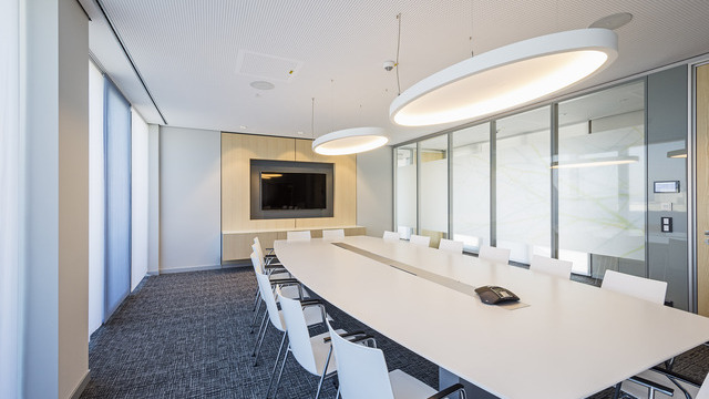 A light-coloured meeting room with a rectangular white table and white chairs in front of a TV screen. Two large elliptical white acoustic lights, SOFT ELLIPSE L+A, are suspended above the table. | © Eckhart Matthäus | www.em-foto.de