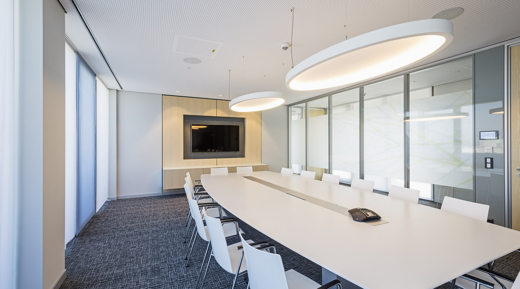 Heller Besprechungsraum mit elliptischer Akustik-Pendelleuchte SOFT ELLIPSE L+A | © Eckhart Matthäus | www.em-foto.de Ein hell gehaltener Besprechungsraum mit einem rechteckigen, weißen Tisch und weißen Stühlen vor einem Fernsehbildschirm. Über dem Tisch sind zwei große elliptische weiße Akustikleuchten, SOFT ELLIPSE L+A, abgependelt. | © Eckhart Matthäus | www.em-foto.de