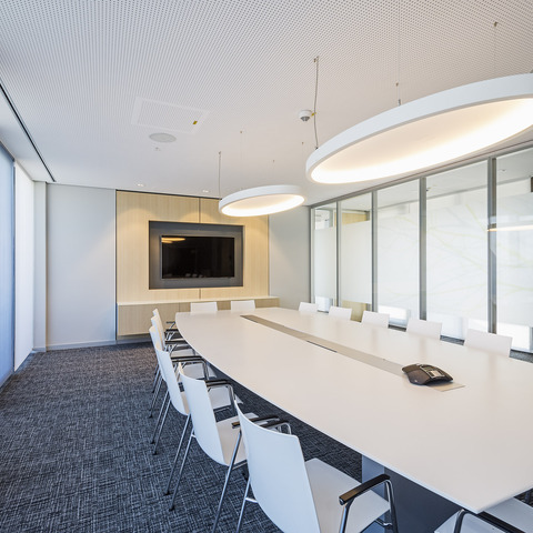 A light-coloured meeting room with a rectangular white table and white chairs in front of a TV screen. Two large elliptical white acoustic lights, SOFT ELLIPSE L+A, are suspended above the table.