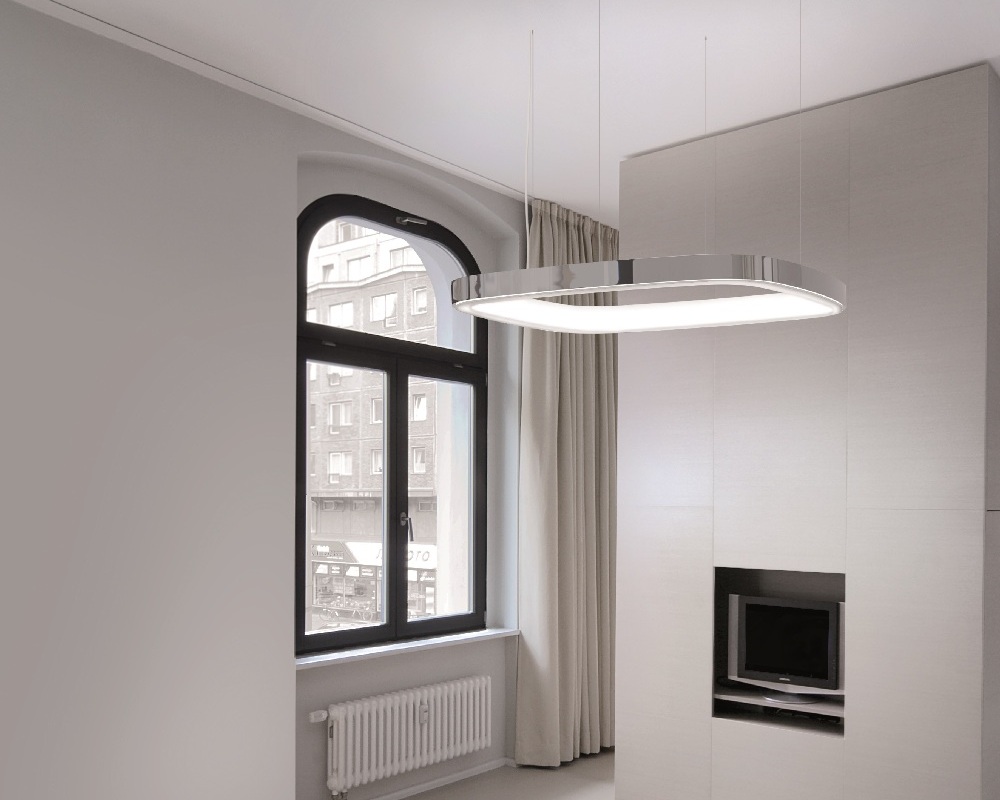 Ring-Pendelleuchte in Form eines abgerundetes Quadrats mit nach unten überstehenden L-Diffusor Große Aluminium-Pendelleuchte in Form eines abgerundeten Quadrats mit L-Diffusor auf der Ringinnenseite, die vor einem Fenster in einem Hotelzimmer abgehangen ist.