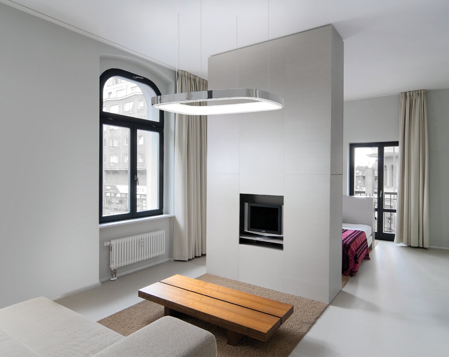 Aluminium-Pendelleuchte in Form eines abgerundeten Quadrats mit L-Diffusor auf der Ringinnenseite, die über einem Holz-Wohnzimmertisch abgehangengen ist, davor steht ein Sofa, dahinter eine Zwischenwand mit Fernseher, hinter dieser sieht man einen Schlafzimmerbereich mit Bett.