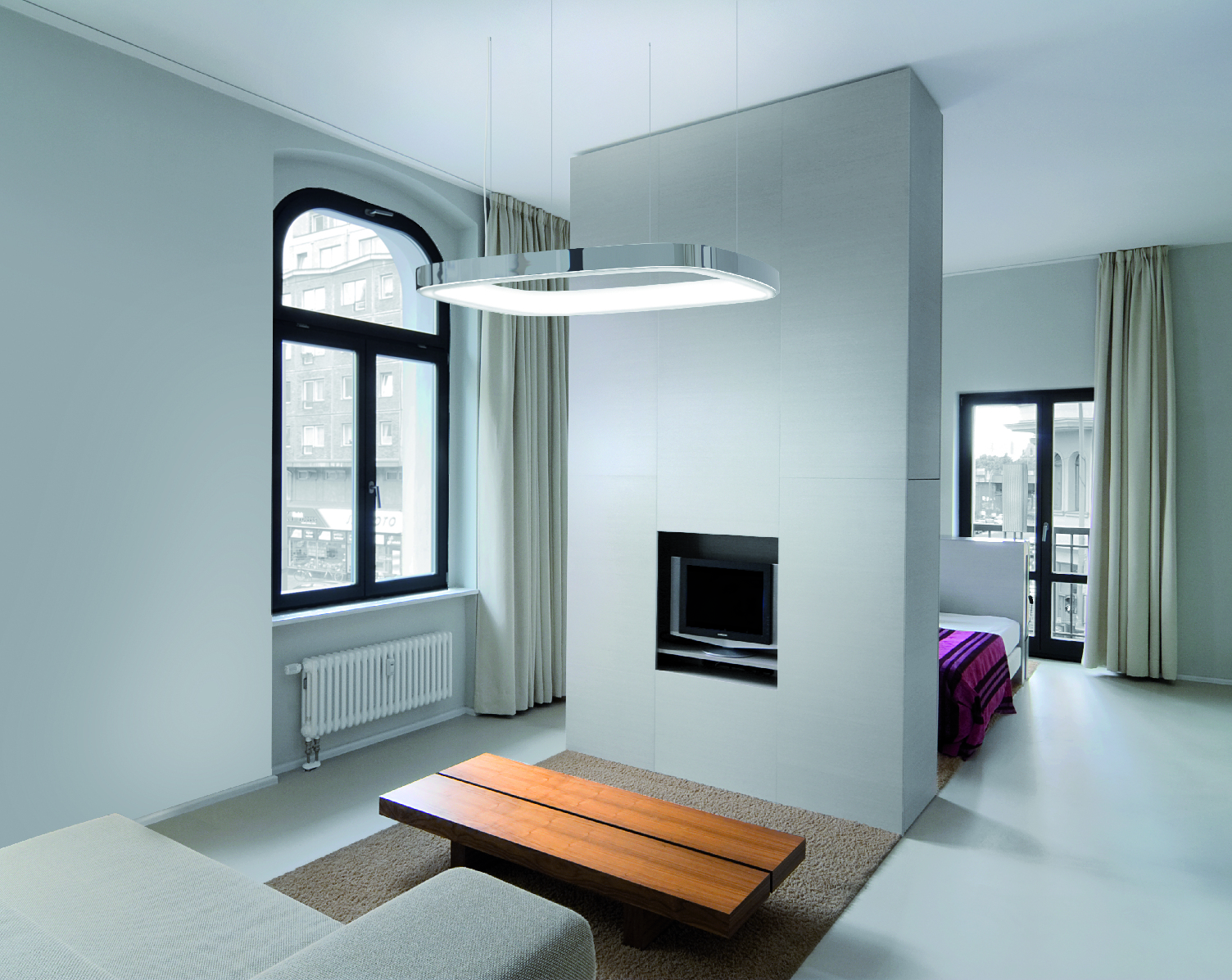 Aluminium-Pendelleuchte in Form eines abgerundeten Quadrats mit L-Diffusor auf der Ringinnenseite, die über einem Holz-Wohnzimmertisch abgehangengen ist, davor steht ein Sofa, dahinter eine Zwischenwand mit Fernseher, hinter dieser sieht man einen Schlafzimmerbereich mit Bett.