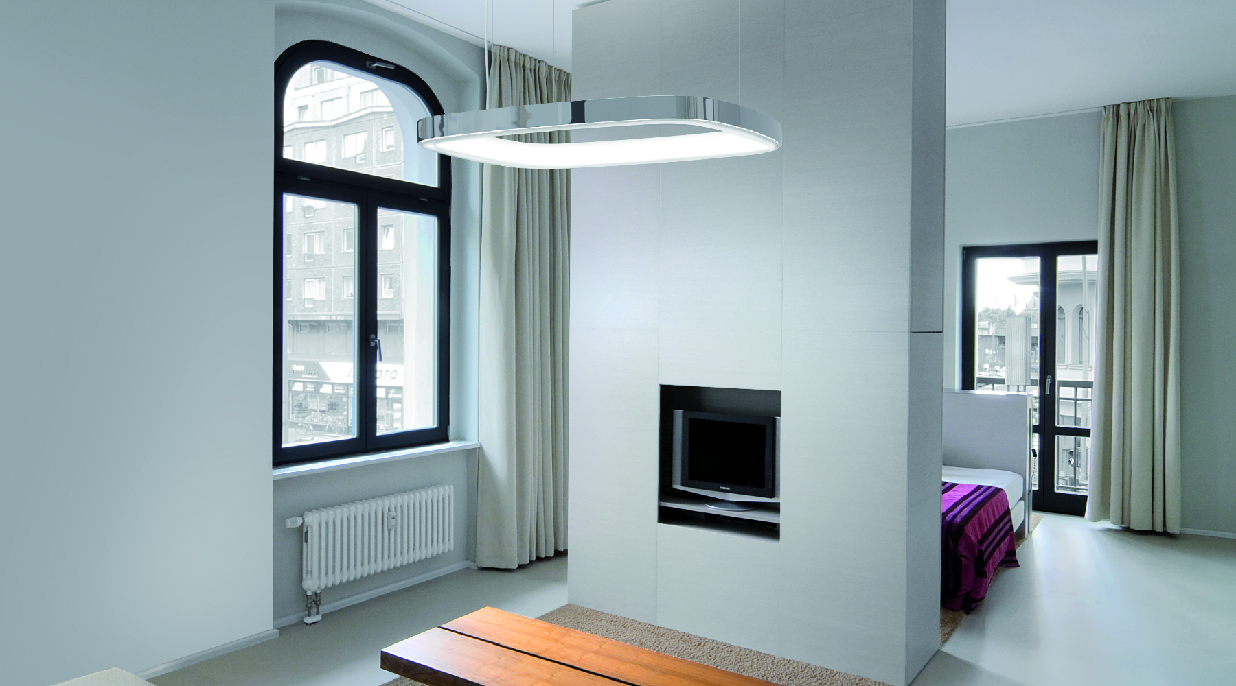 SOFT QUADRA - abgerundetes Quadrat leuchtet über Wohnzimmertisch Aluminium-Pendelleuchte in Form eines abgerundeten Quadrats mit L-Diffusor auf der Ringinnenseite, die über einem Holz-Wohnzimmertisch abgehangengen ist, davor steht ein Sofa, dahinter eine Zwischenwand mit Fernseher, hinter dieser sieht man einen Schlafzimmerbereich mit Bett.