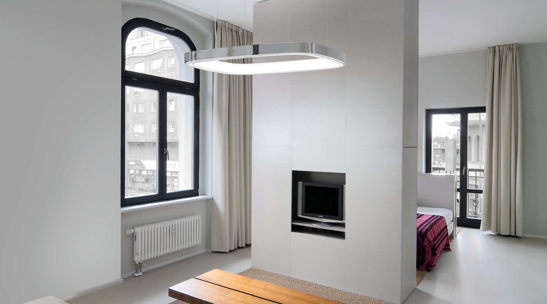 SOFT QUADRA - abgerundetes Quadrat leuchtet über Wohnzimmertisch Aluminium-Pendelleuchte in Form eines abgerundeten Quadrats mit L-Diffusor auf der Ringinnenseite, die über einem Holz-Wohnzimmertisch abgehangengen ist, davor steht ein Sofa, dahinter eine Zwischenwand mit Fernseher, hinter dieser sieht man einen Schlafzimmerbereich mit Bett.