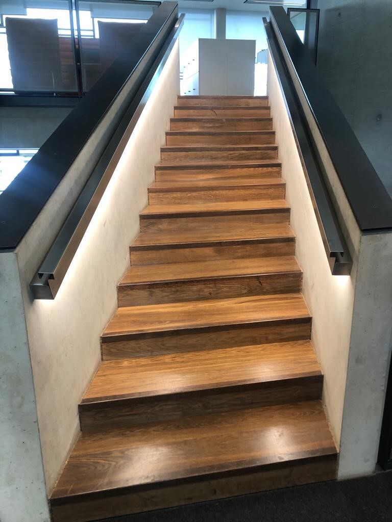 Straight staircase with wooden treads, concrete side walls, a metal handrail on the inside of the walls on both sides, from which light shines downwards onto the steps.