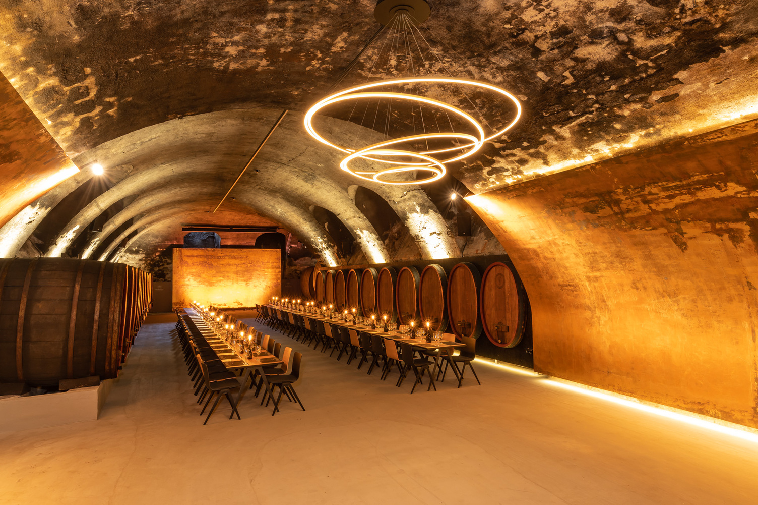 Vaulted wine cellar of the Staatliche Hofkellerei Würzburg with wooden barrels along the walls, in the centre is a long, festive table consisting of several wooden tables and chairs with candles on the tables, above which hovers a modern chandelier consisting of several interconnected ring lights (TOCCATA). | © Staatlicher Hofkeller Würzburg | Photo: Brigitte Sauer