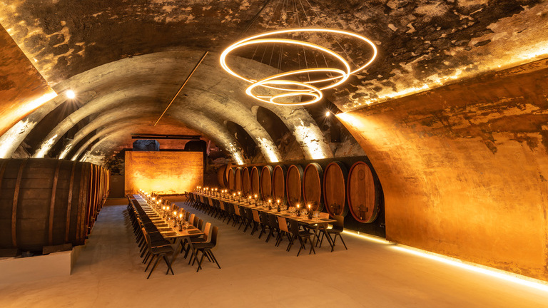 Vaulted wine cellar of the Staatliche Hofkellerei Würzburg with wooden barrels along the walls, in the centre is a long, festive table consisting of several wooden tables and chairs with candles on the tables, above which hovers a modern chandelier consisting of several interconnected ring lights (TOCCATA). | © Staatlicher Hofkeller Würzburg | Photo: Brigitte Sauer