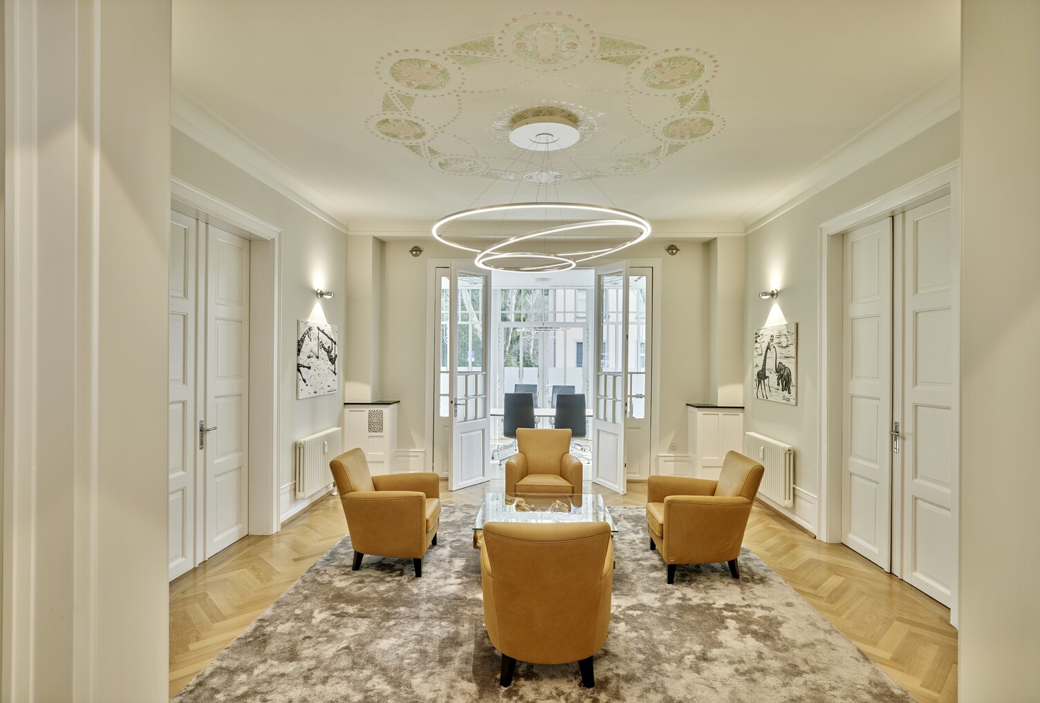 Waiting area in a classic office villa with 4 yellow armchairs around a glass table, above the glass table hovers a modern ring chandelier (TOCCATA) suspended from an ornate ceiling. | © Christoph Tempes