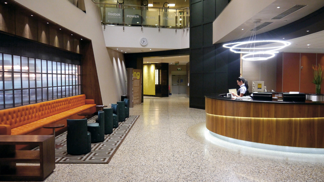 Large reception area with a lounge area on the left of the picture, opposite a round wooden reception desk, a modern chandelier in the form of connected ring lights, TOCCATA, is suspended above the reception desk.