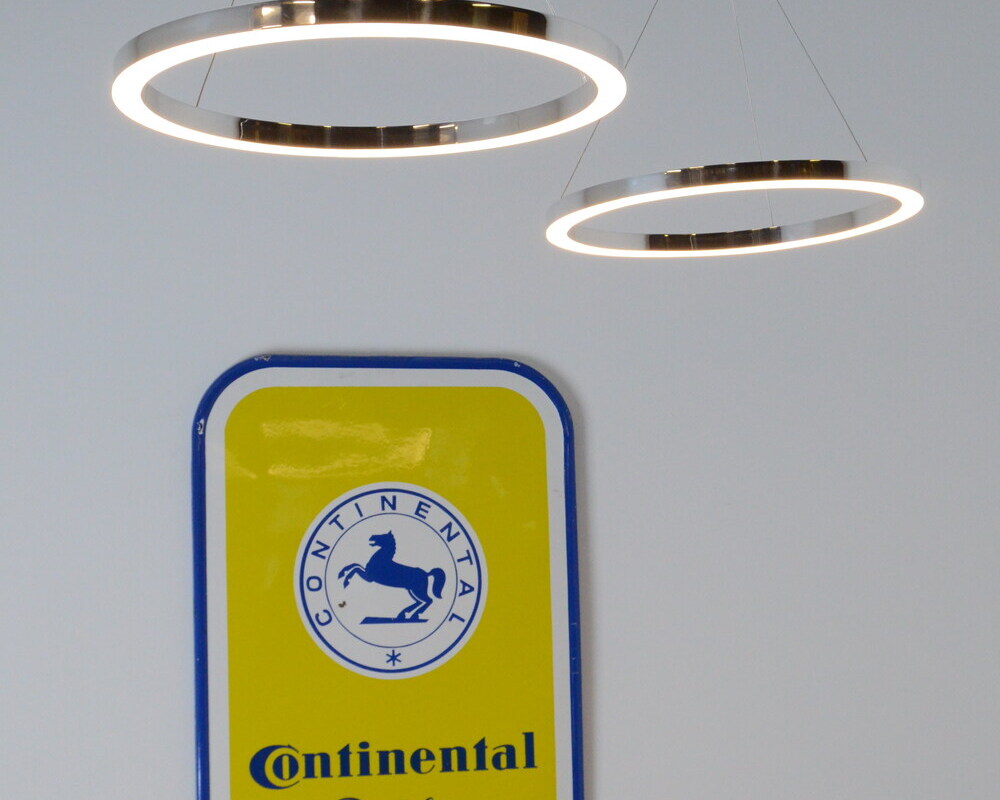 Zwei kleine polierte Aluminium Ringleuchten auf unterschiedlicher Höhe vor einem Metallschild von Continentale abgependelt Im Winkel kippbar. TRIS Pendelleuchte.