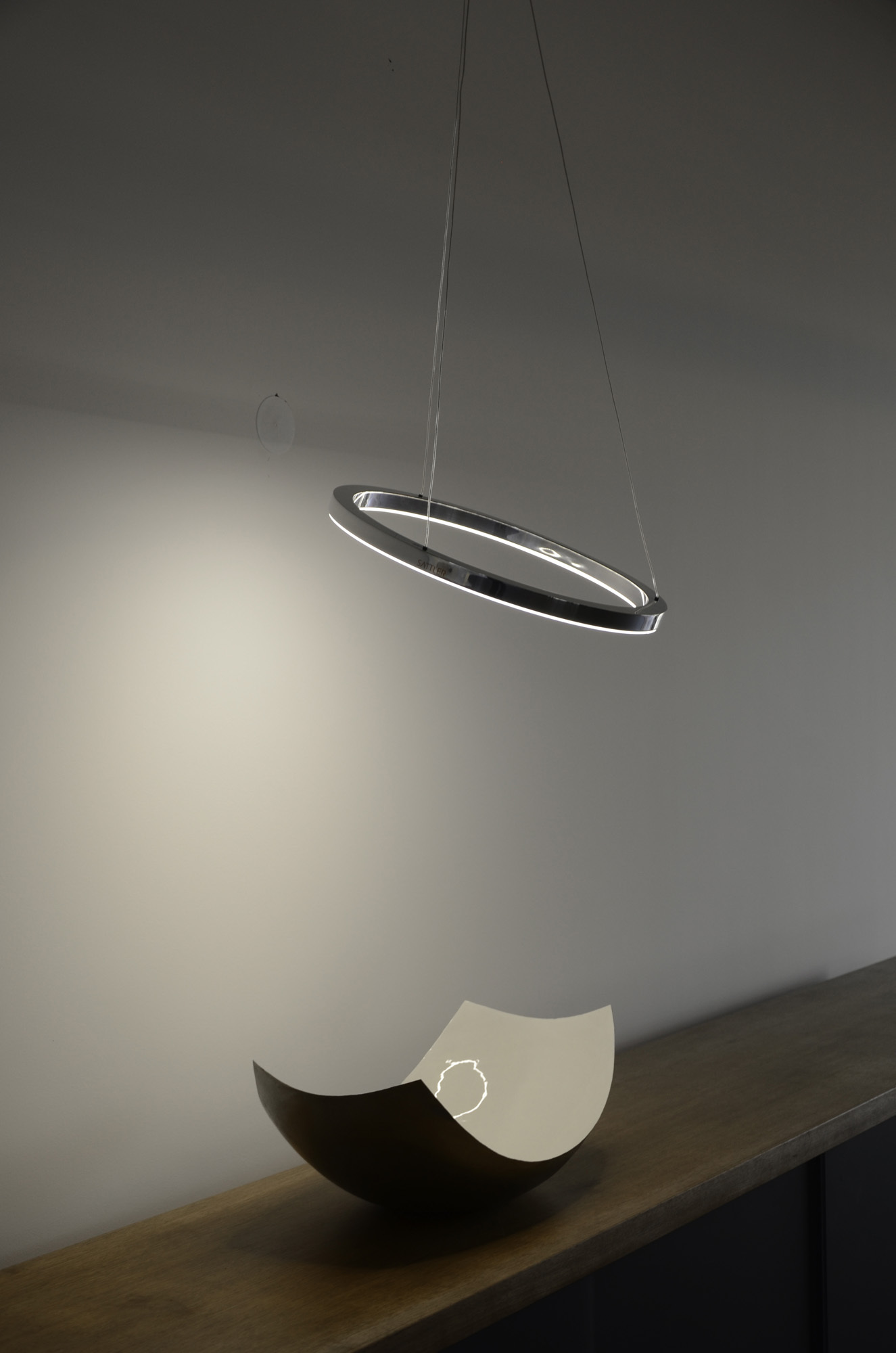 TRIS: Aluminium-polierte Ringleuchte, leicht gekippt abgependelt, darunter eine moderne Metallschale auf Holz-Sideboard.
