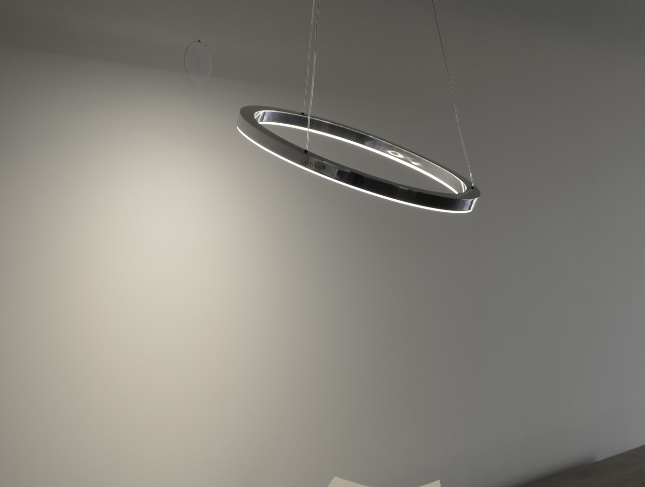 TRIS: Aluminium-polierte Ringleuchte, leicht gekippt abgependelt, darunter eine moderne Metallschale auf Holz-Sideboard.