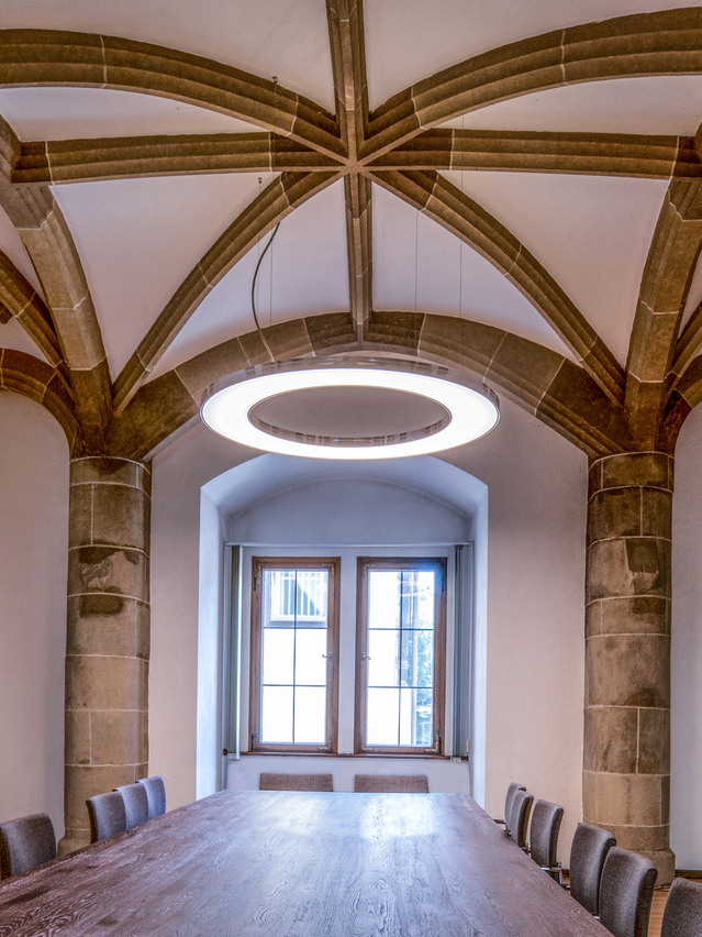 Großer Gerichtssaal im Amtsgericht Göppingen mit einem großen Holztisch und Stühlen darum. Die Decke ist eine historische Gewölbedecke mit Steinernen Verstrebungen. Von dieser ist eine großflächige Ringleuchte, ANELLO, abgependelt. | © SATTLER
