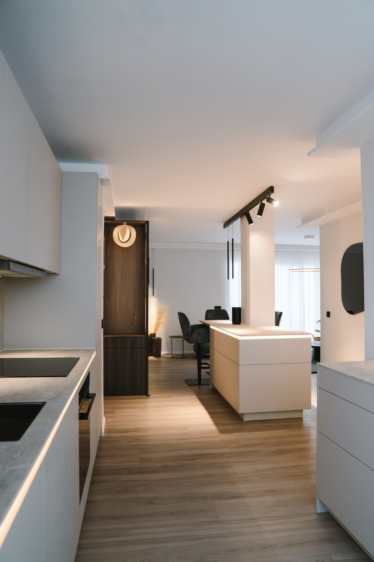 Blick in einen modernen Flur entlang dessen eine moderne Küchenzeile verläuft. Am Ende des Flurs links ein Schrank an dem ein Hut aufgehängt ist, rechts daneben ein Blocktisch mit einer linearen Leuchte, bzw. PALADIO TRACK Stromschiene darüber, in der zylindrische EVOLUTION SPOT Leuchten und zwei zylindrische FINO PENDANT Hängelampen eingefügt sind, die den Hochtisch beleuchten. | © Pia Schöning
