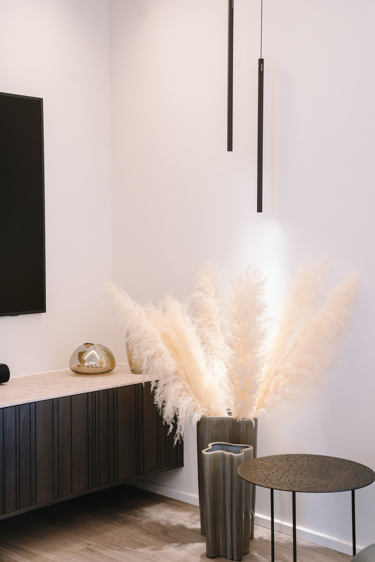 Dekorative Wohnzimmerleuchten FINO PENDANT Dekorative Wohnzimmerecke mit Ausschnitt von einem Sideboard und Fernseher darüber, das Eck wird ausgeglichen durch eine Vase mit Grasartigen Pflanzen. Diese werden beleuchtet von zwei schmalen zylindrischen Pendelleuchten, FINO PENDANT, die in unterschiedlichen Höhen darüber abgehangen sind.
