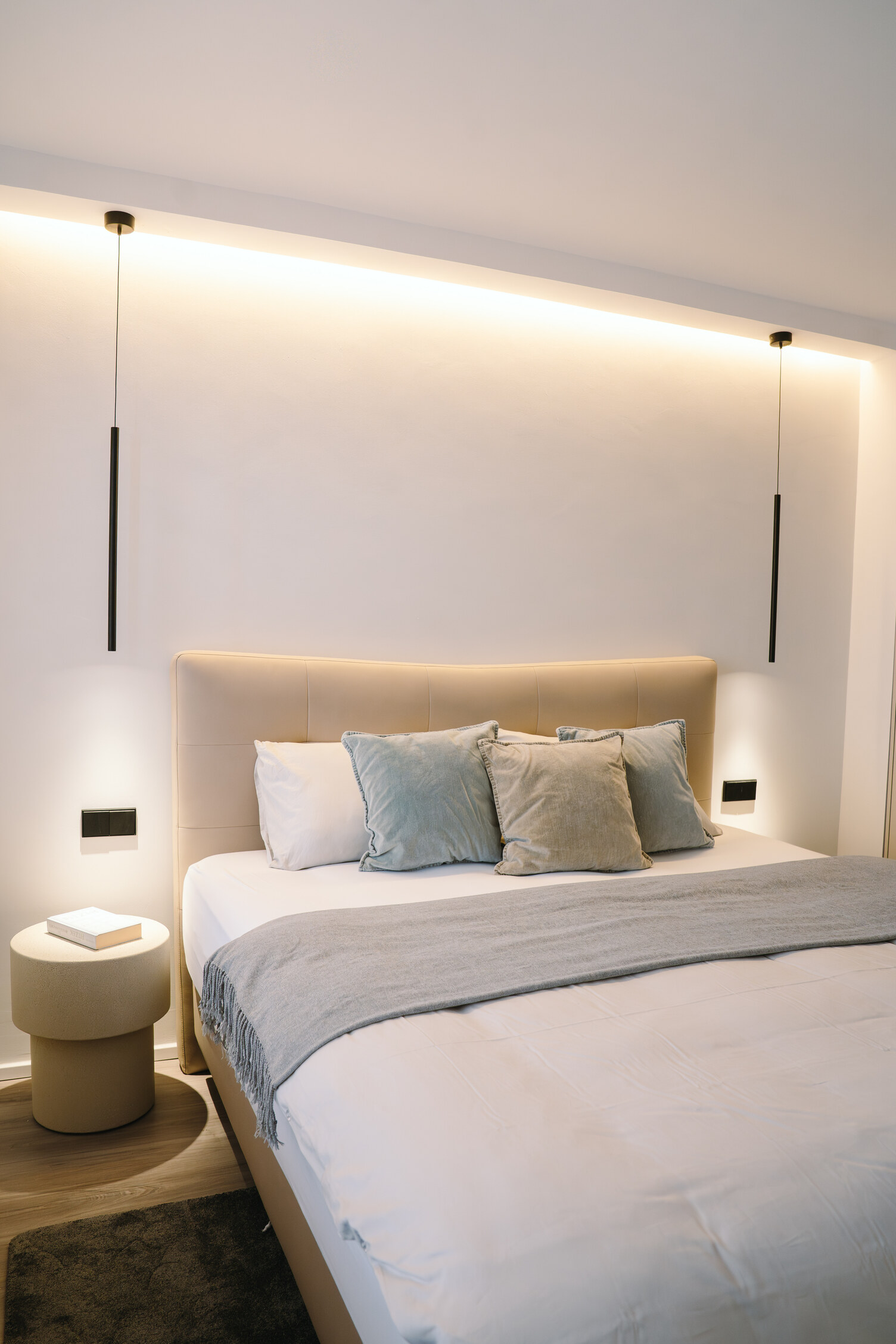Elegantes Ambientelicht Schlafzimmer Modernes helles Doppelbett mit bunten Kissen in Pastelltönen. Neben dem Bett kleine runde Beistelltische, die von einer zylindrischen Hängelampe darüber, FINO PENDANT, angestrahlt werden.
