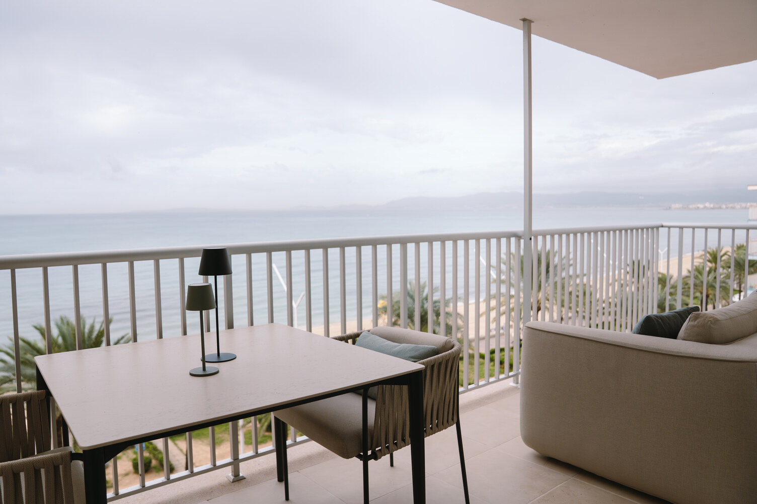 Balkonblick vom Apartment Arenal auf das Meer Blick vom Rauminneren auf einen Balkon am Meer. Man sieht einen Tisch mit zwei Stühlen und einem Außensofa. Das weiße Geländer ist unterbrochen und eröffnet den Blick auf Strand und Meer. Arenal Appartment in Mallorca. Innenarchitektur und Einrichtung: INSIGHT Design Mallorca