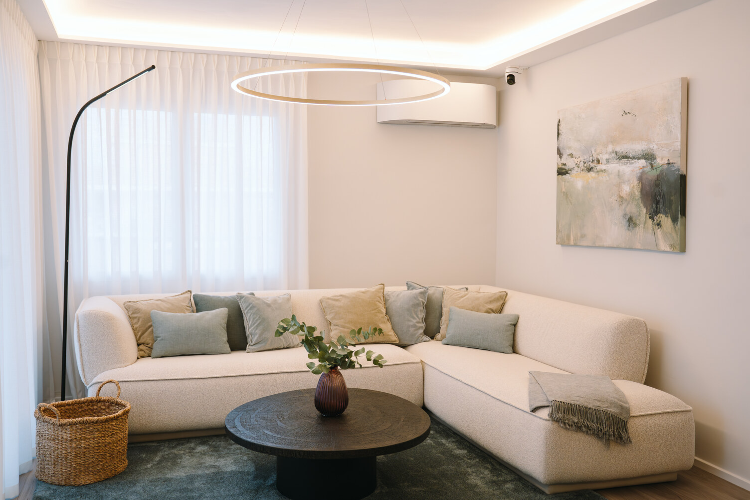 Modernes Wohnzimmer mit einem blauen Teppichboden. Den Mittelpunkt des Raumes bildet ein helles Sofa mit pastellfarbenen Kissen. Das Zentrum wird markiert durch eine dekorative Wohnzimmerlampe, der Ringleuchte SLIM DIRECT. Weiteres dekoratives und Lichtvolles Element, das über dem Sofa gebogen hervorragt ist die Wohnzimmerstehleuchte PALITO FLOOR, die an eine Peitschenleuchte erinnert. Rechts an der Wand hängt ein schwarz weiß Bild. 