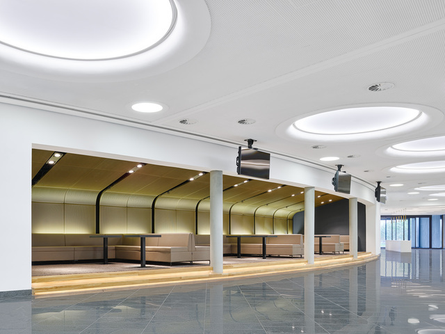 Runde Lichtdecken. Loungebereich in Goldfaben mit Sitzbänken als Nischen in einer großen Halle mit weißer Decke in der große rund Lichtfelder angeordnet sind. | © Zooey Braun