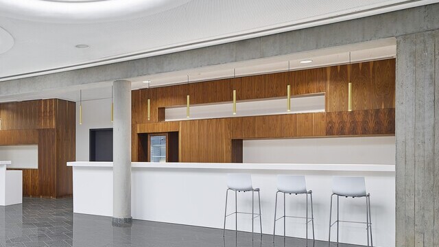 Zoom on counter in a large hall with stone floor and a white ceiling in which large round light fields are arranged, view goes into a counter area, which is characterised as niches and is highlighted by brown wooden walls, in addition, golden bar lights (TIAs) are suspended at regular intervals over the entire counter. | © Zooey Braun