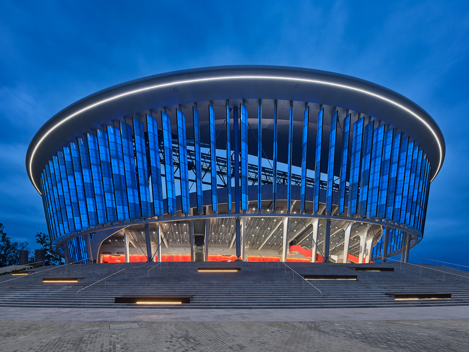 VIP Lounge mit SATTLER Beleuchtung. Außenansicht eines modernen runden Veranstaltungsstadions, der Black Sea Arena in Georgien, das  von einem hellen Lichtband oben umrandet ist und darunter in blau erleuchtet ist. | © Zooey Braun