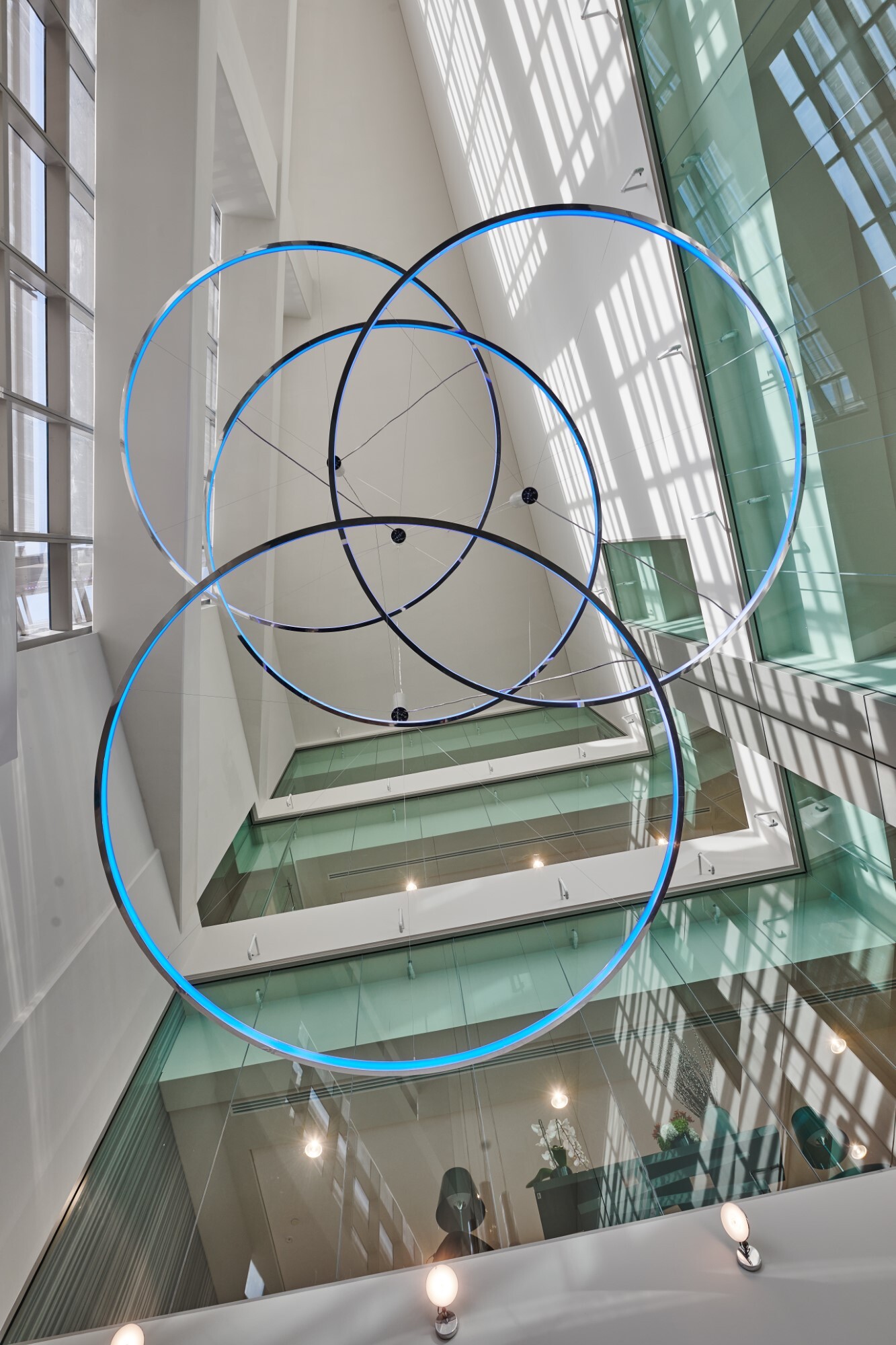 Blick von unten in einem Glasatrium auf 4 x Ringleuchte CIRCOLO-I mit Pendelbaldachin, Ø 3500mm, Gehäuse aus Aluminium poliert und hellblau leuchtend, an den Wänden 12 x AVVENI-Single Spots.