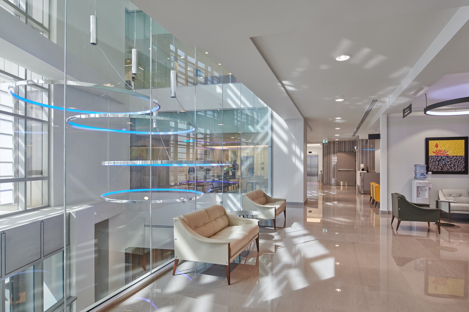 Hospital corridor, left in the picture View of a glass atrium with four large polished aluminium ring luminaires CIRCOLO-I with pendant canopy, which shine in blue and are suspended at different heights.