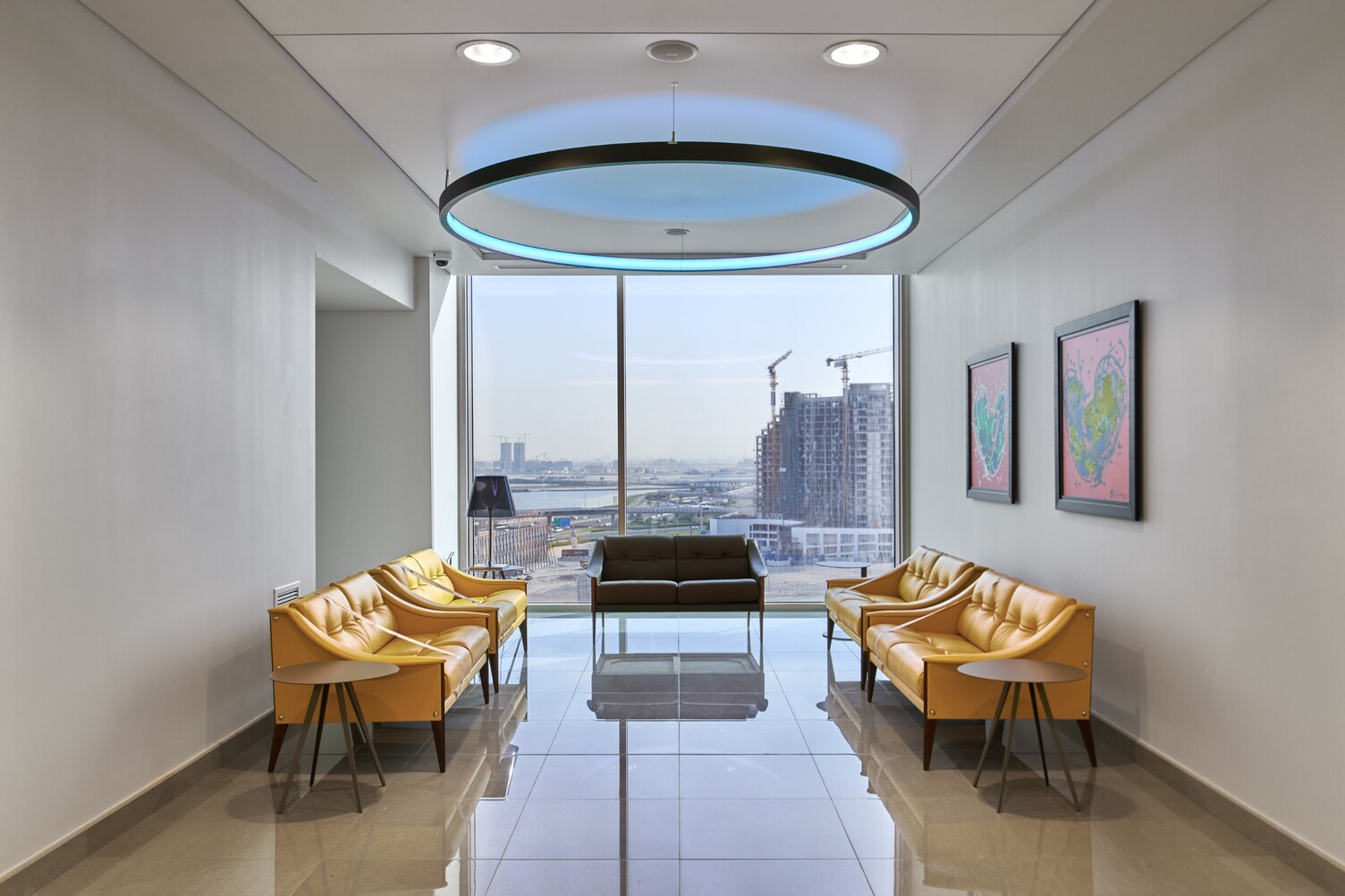 View into a waiting area with a large window front and a view of Dubai's skyscrapers; a round black pendant ring light can be seen shining blue above the furniture in the centre of the area.