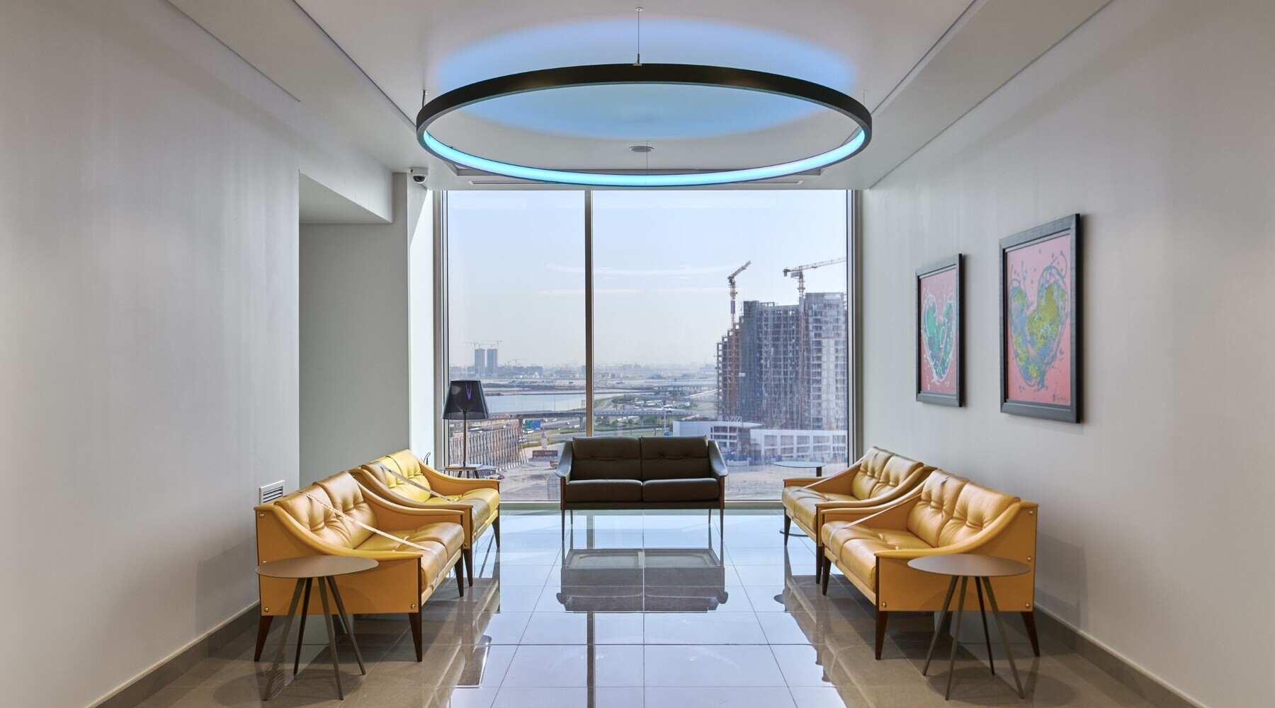 © © LUMIERE GROUP View into a waiting area with a large window front and a view of Dubai's skyscrapers; a round black pendant ring light can be seen shining blue above the furniture in the centre of the area. | © © LUMIERE GROUP