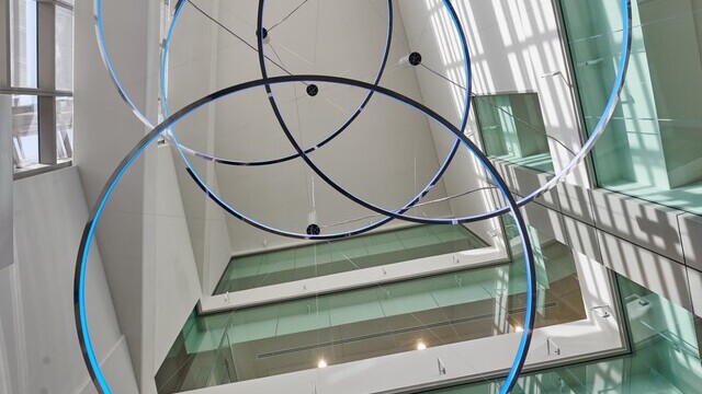 Blick von unten in einem Glasatrium auf 4 x Ringleuchte CIRCOLO-I mit Pendelbaldachin, Ø 3500mm, Gehäuse aus Aluminium poliert und hellblau leuchtend, an den Wänden 12 x AVVENI-Single Spots. | © © LUMIERE GROUP