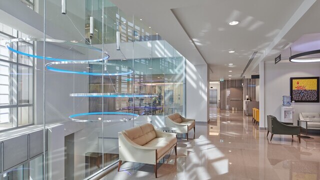 Hospital corridor, left in the picture View of a glass atrium with four large polished aluminium ring luminaires CIRCOLO-I with pendant canopy, which shine in blue and are suspended at different heights. | © LUMIERE GROUP