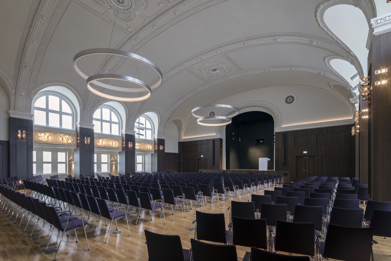 A hall in the historic Besenbinderhof building in Hamburg, featuring rows of purple chairs; suspended from the stuccoed vaulted ceiling, two sets of two large CIRCOLO ring lights are arranged one above the other along the length of the room, resembling a small, modern chandelier.  | © Michael Voit