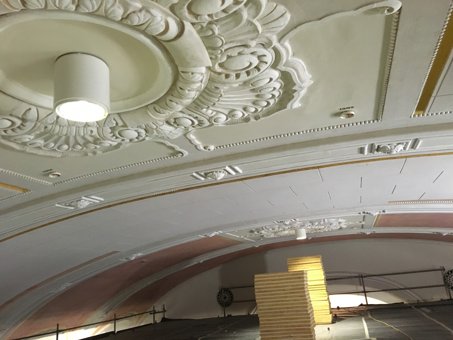 A mezzanine level with scaffolding extending into the room. Close to the vaulted ceiling, which is decorated with stucco rosettes. Simple cylindrical spotlights are fitted within the stucco rosettes.