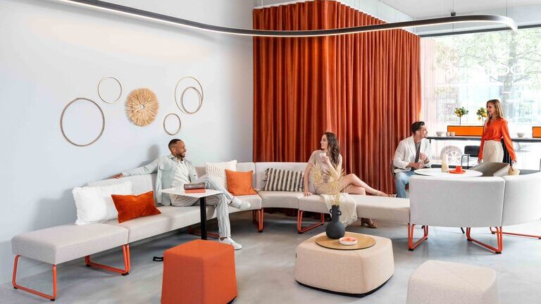 Stylish lounge area in a modern showroom with orange accents and Sattler's Duetto curved lighting fixture in Offenbach. | ©  Dauphin HumanDesign® Group GmbH & Co. KG