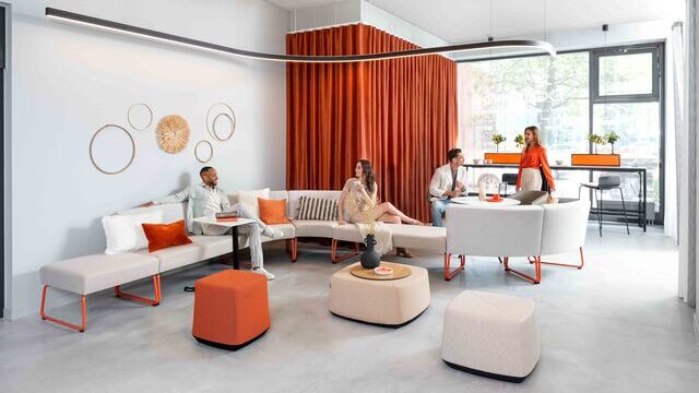 Stilvolle Lounge-Bereich in einem modernen Showroom mit orangenen Akzenten und Sattlers Duetto gebogenem Leuchtkörper in Offenbach. | ©  Dauphin HumanDesign® Group GmbH & Co. KG