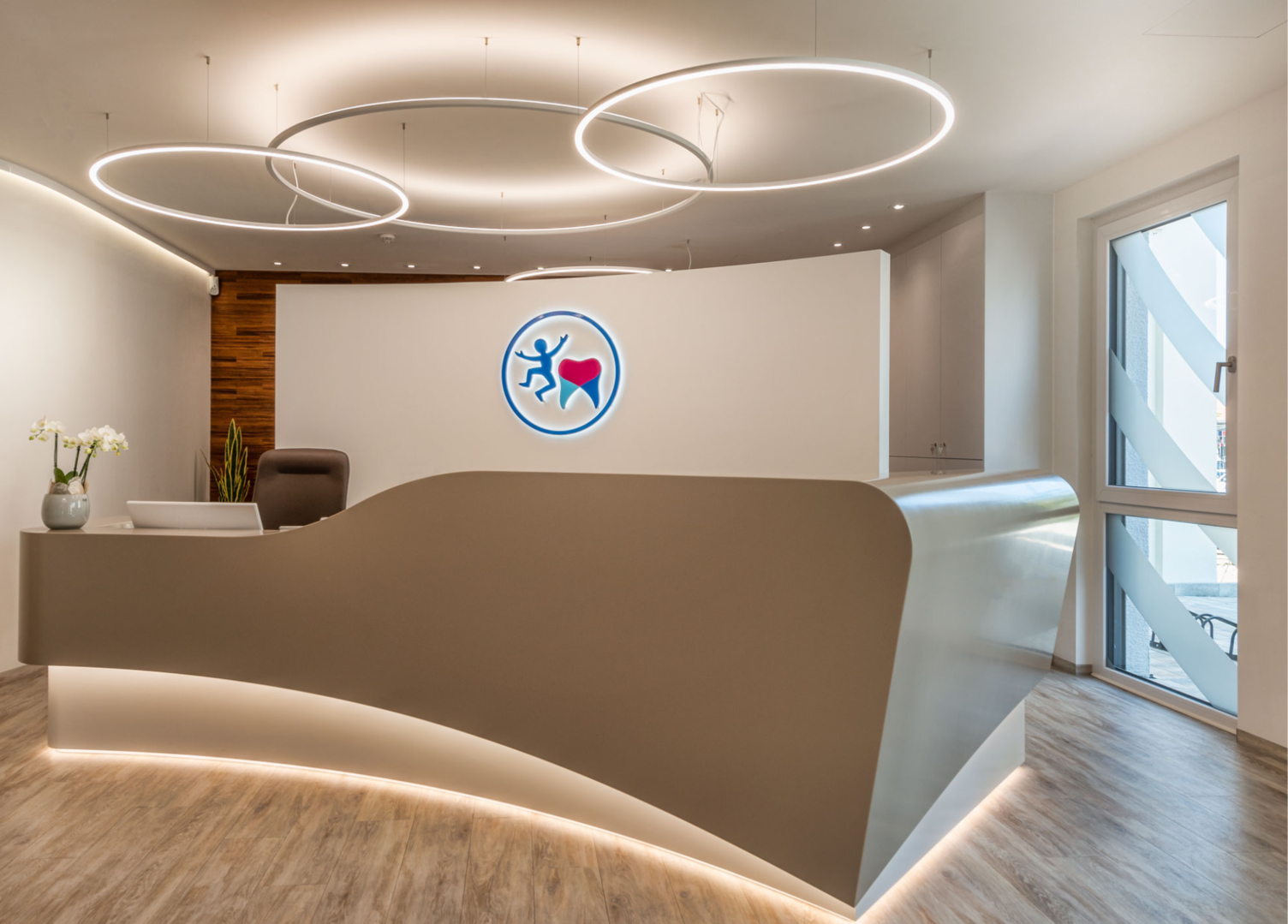 Beige geschwungene Empfangstheke Kieferorthopädiepraxis Dr. Grammatidis, bodennah abgesetzt und hinterleuchtet, auf der Rückwand ist ein Zahn und ein Strichmännchen, über der Theke hängen 3 schmale SLIM Ringleuchten von SATTLER versetzt angeordnet, zwei leuchten direkt, eine leuchtet indirekt, und hellt die Decke auf.