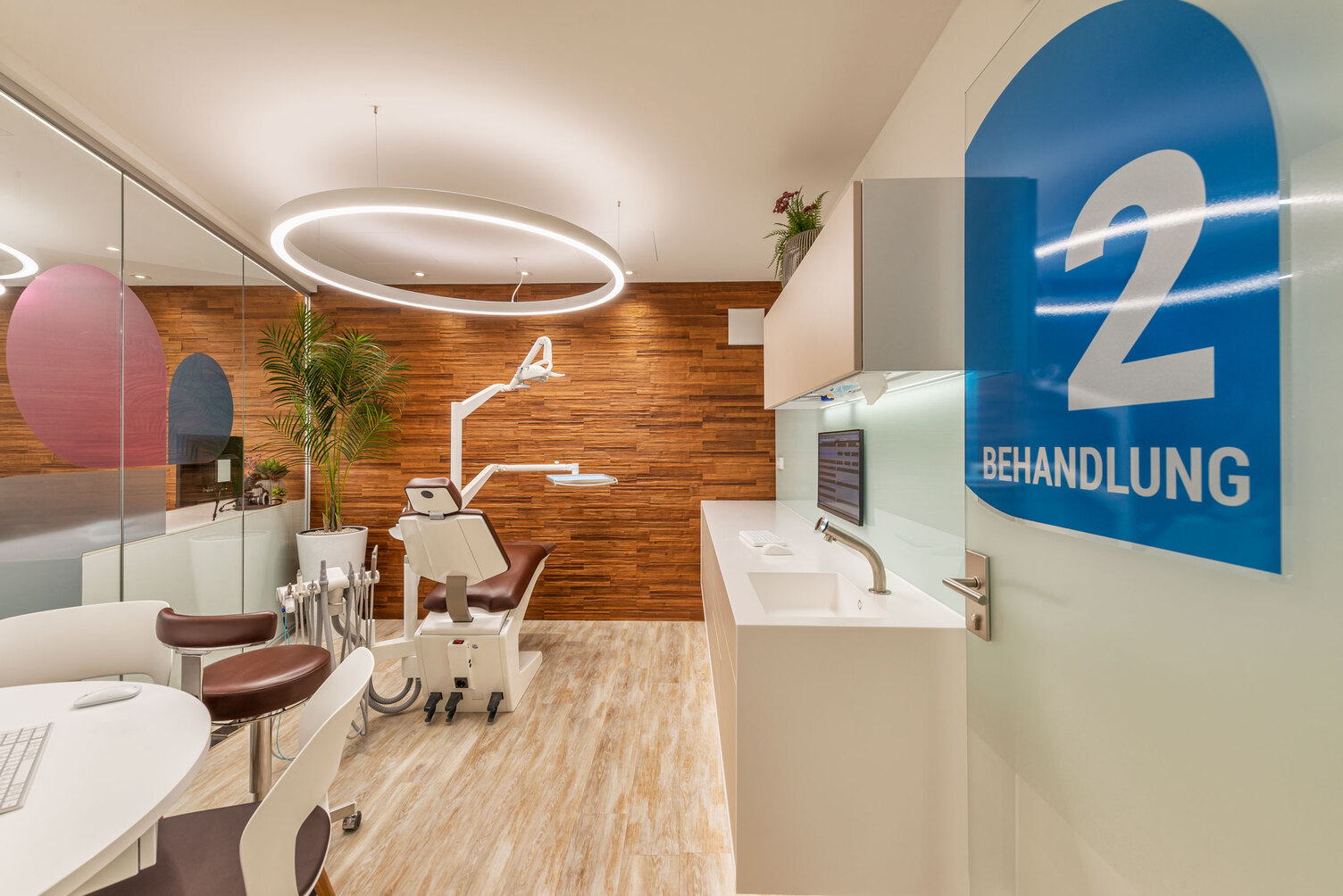 Modern, warm treatment room at Dr Grammatidis, Kirchheim; specialist dentists for orthodontics, with focus on a dental treatment chair above which a white DUETTO MED ring light is suspended.