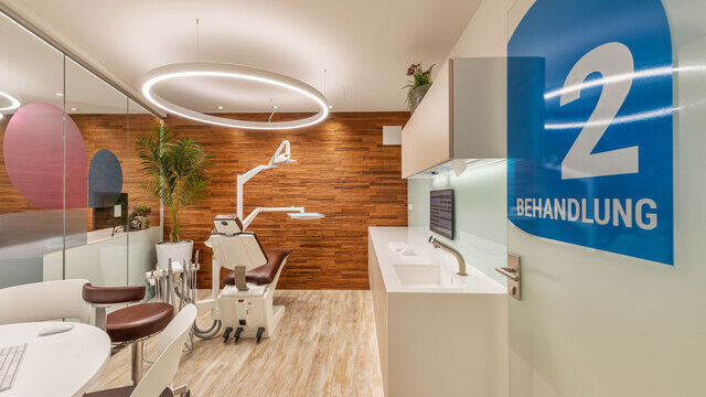 Modern, warm treatment room at Dr Grammatidis, Kirchheim; specialist dentists for orthodontics, with focus on a dental treatment chair above which a white DUETTO MED ring light is suspended. | © faktorzwei GmbH