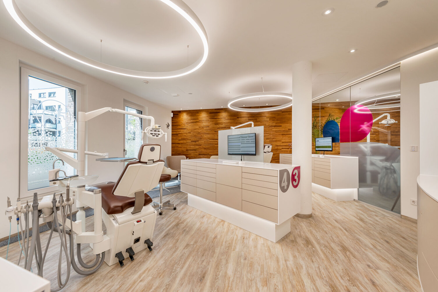 Dental practice Dr Grammatidis with several treatment chairs above which a ring light is suspended, pleasant atmosphere also due to the brown structured wooden wall.