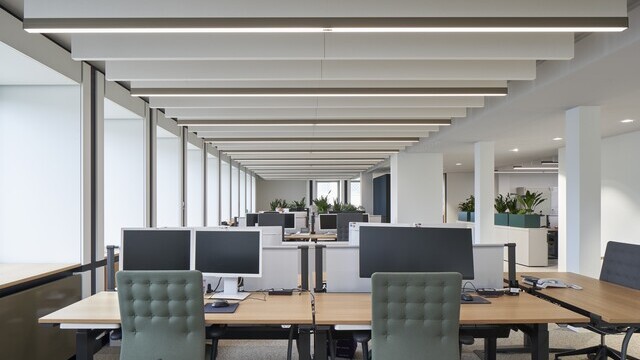Blick in ein Großraumbüro mit unendlich wirkender Raum mit Bildschirmarbeitsplätzen, grünen Bürostühlen und an der Decke lineare Leuchten und weißen Akustikbaffeln.