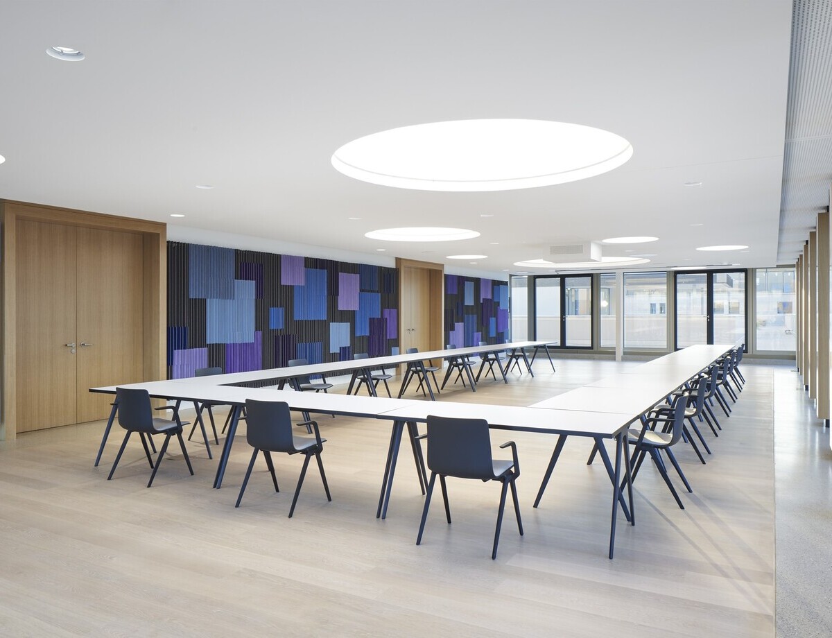 Konferenzraum mit einer großen U-Anordnung der Tische, die Wände sind in Blautönen grafisch gestaltet,  in der weißen Decke sind runde Lichtdecken im Raum verteilt.
