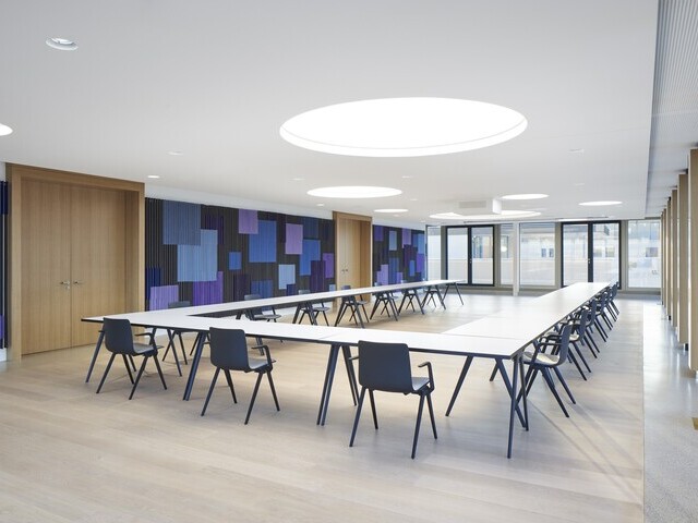 Konferenzraum mit einer großen U-Anordnung der Tische, die Wände sind in Blautönen grafisch gestaltet,  in der weißen Decke sind runde Lichtdecken im Raum verteilt.