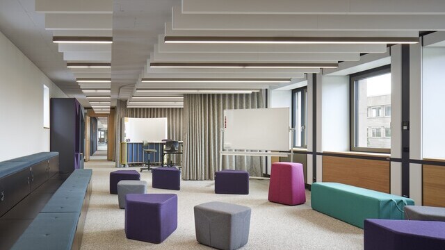 Workshop office space with colourful upholstered furniture and seating cubes, above which several linear acoustic baffles and lights can be seen in two rows one behind the other. | © Constantin Meyer